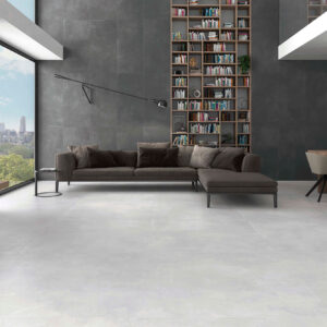 AMB-ELEMENTI-GRAPHITE-60x120+-PEARL-120x120-HQ2-CMYK Sala de exposición con pavimento cerámico gris claro y revestimiento mural oscuro, sofá y comedor