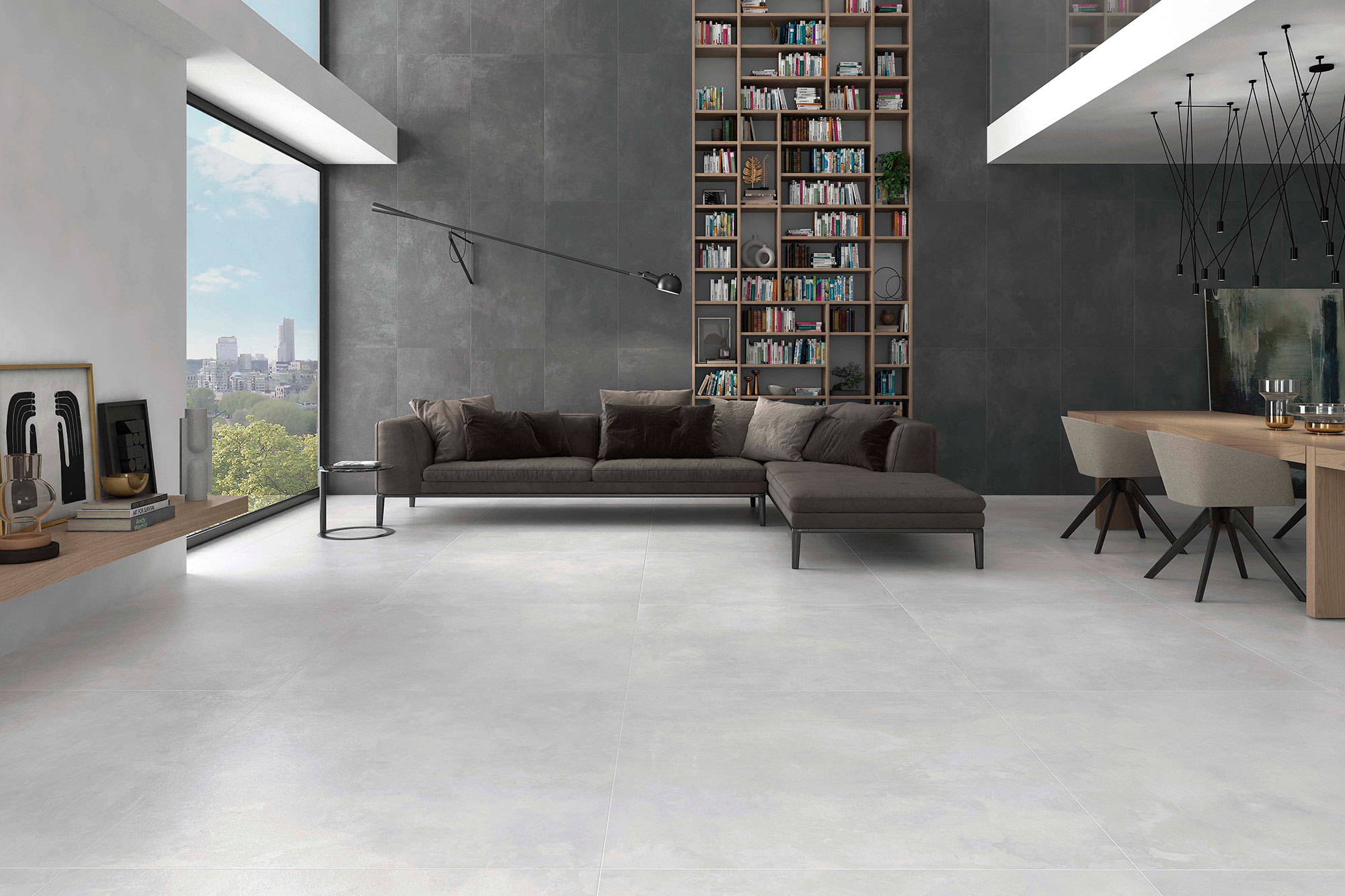 AMB-ELEMENTI-GRAPHITE-60x120+-PEARL-120x120-HQ2-CMYK Sala de exposición con pavimento cerámico gris claro y revestimiento mural oscuro, sofá y comedor