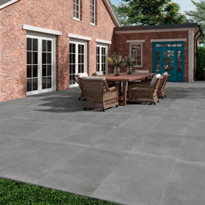 AMB-SONDER-ANTRACITE-60X120-20MM Terraza exterior con pavimento cerámico gris y zona de comedor junto a casa de ladrillo.