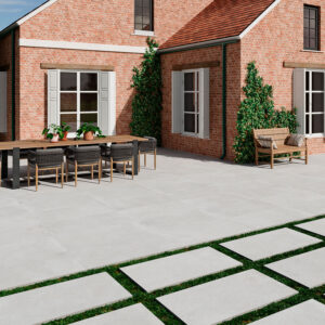 AMB-SONDER-PEARL-60X120-20MM Patio con pavimento cerámico gris, losas con franjas de césped y mesa junto a fachada de ladrillo.