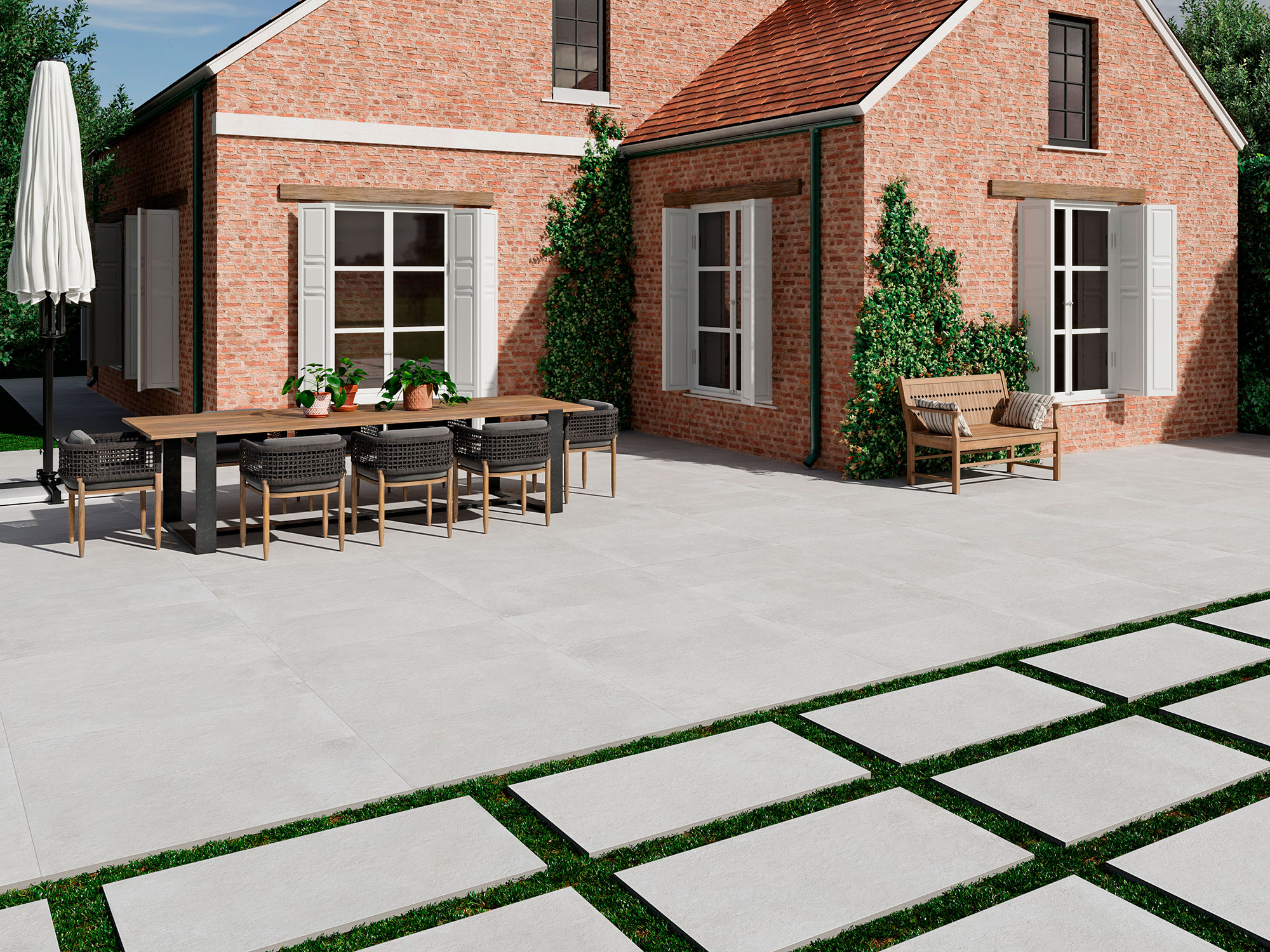 AMB-SONDER-PEARL-60X120-20MM Patio con pavimento cerámico gris, losas con franjas de césped y mesa junto a fachada de ladrillo.