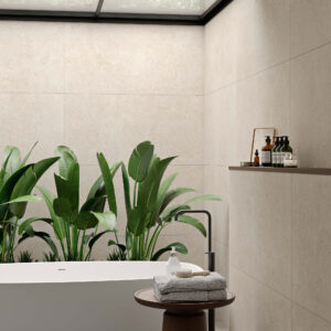 Baño con bañera y azulejos cerámicos beige, plantas y estante con accesorios