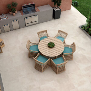 Patio exterior con mesa redonda de madera, seis sillas de mimbre y pavimento cerámico beige.