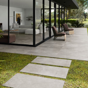Terraza con grandes losas cerámicas grises, camino en placas sobre césped y salón acristalado.