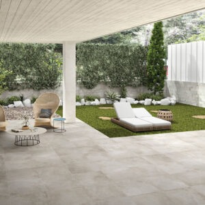 Terraza cubierta con amplias losas cerámicas grises, sillones de mimbre y tumbona en césped