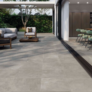 STN-AMB-ELEMENTI-GREY-45X45-INOUT Terraza y comedor abiertos con pavimento cerámico gris, sofás, mesa y jardín.