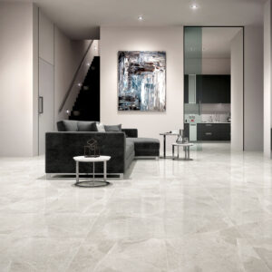 0815_ALAPLANA_chelsea-gris Salón moderno con sofá oscuro, cocina abierta y suelos cerámicos gris claro pulidos