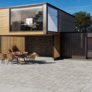 Patio con pavimento cerámico gris, adoquinado central y muebles exteriores frente a vivienda moderna