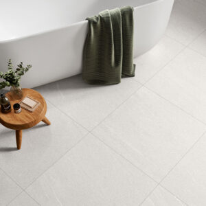 AMB-HALTON-WHITE-60X60-DETALLE Baño con bañera blanca sobre pavimento cerámico gris claro, mesa de madera y toalla verde.